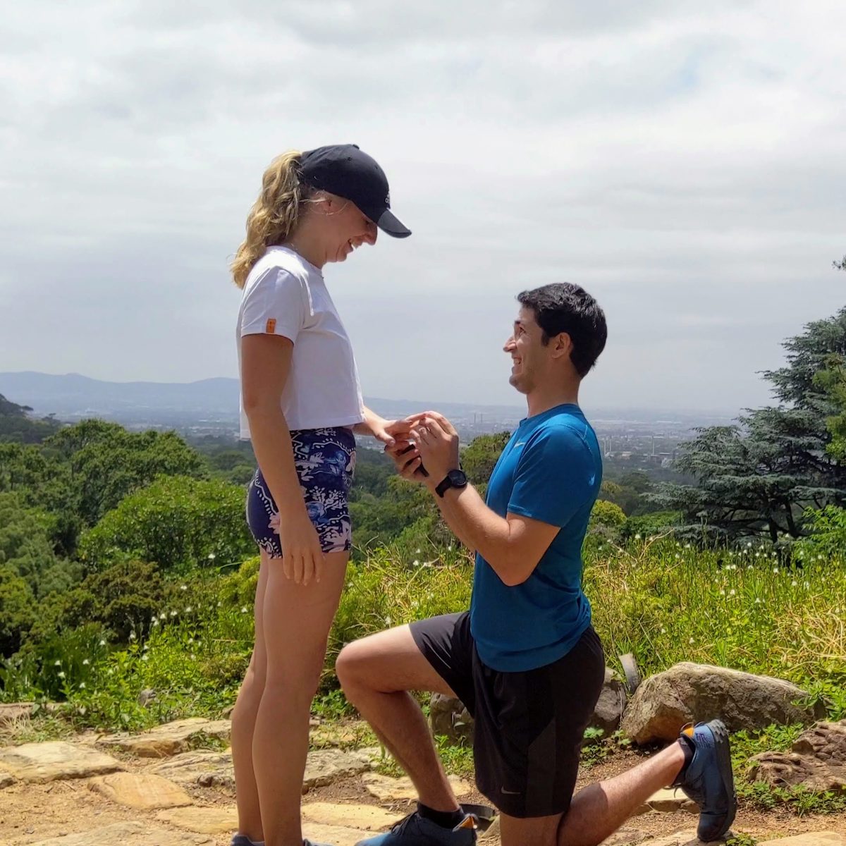 Wesley on one knee, proposing to Claire on a mountainside lookout above Cape Town